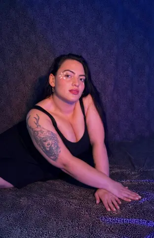 Curvy woman in a black tank top relaxing on a textured dark sofa under moody blue lighting.