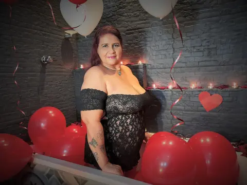 LaurenRiley poses in a black lace off-the-shoulder dress, surrounded by red and white balloons against a dark brick wall.