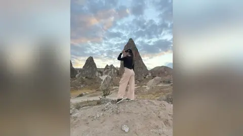 Stylish woman in black top and beige trousers standing against a dramatic mountain landscape at sunset.