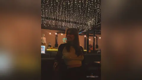 Young woman in a beige top standing under festive fairy lights in a dimly lit lounge setting.