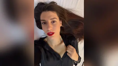 Young woman with red lipstick wearing a black silk blouse lying on white bedding with a relaxed mood.