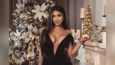 Glamorous woman in a black evening dress posing by a decorated Christmas tree and festive mantelpiece.