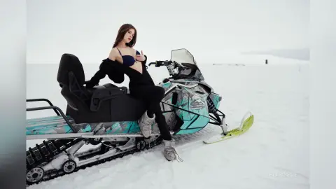 Woman in black winter outfit sitting on a teal snowmobile in a vast snowy landscape.