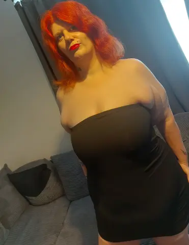 leeanneHill poses in a black strapless mini dress, showing her red hair and cleavage while leaning back against a grey sofa.