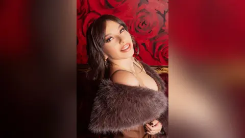 Smiling brunette woman in a brown faux fur coat posing against a romantic red rose floral background.