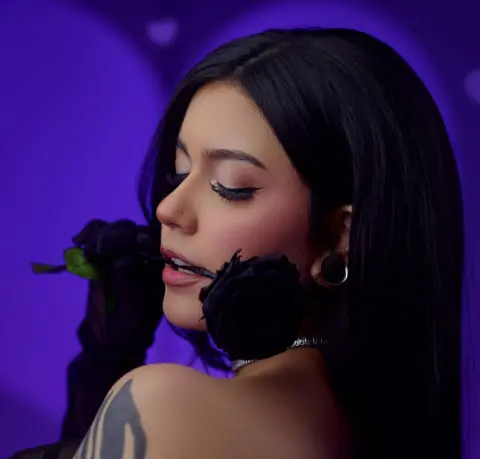 Glamorous woman with long black hair and dark makeup posing with black roses against a purple background.
