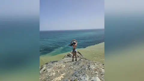 RossiNoleen stands on a rocky cliff overlooking a clear blue ocean, wearing light blue denim shorts.