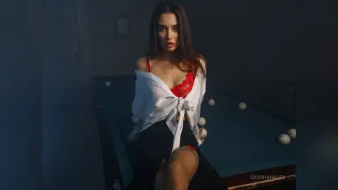 Young woman in a white shirt and red lace lingerie sitting by a pool table in a moody, dimly lit room.