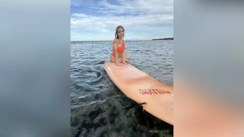 Smiling woman in an orange swimsuit sitting on a surfboard in the ocean under a bright blue sky.