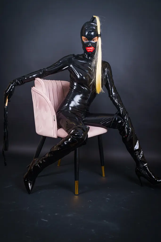AlexaDuque kneels on a dark surface, wearing a black bra and posing with her head down, showing her bare back and legs.