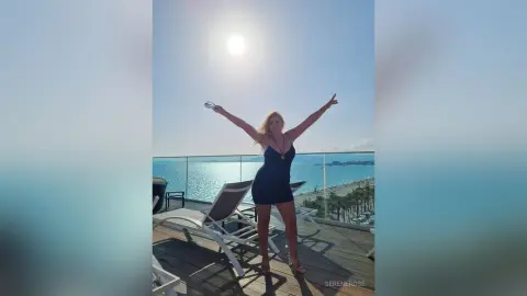 Cheerful woman in a black cocktail dress raising arms on a sunny tropical resort balcony overlooking the ocean.