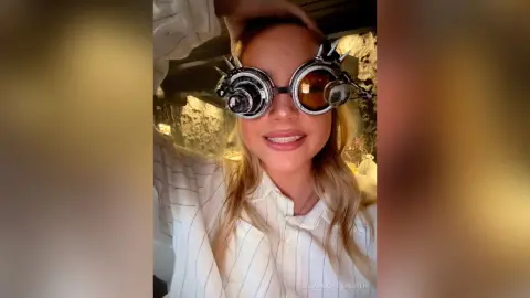 Smiling blonde woman in a white striped shirt wearing oversized steampunk goggles in a warm, dimly lit setting.