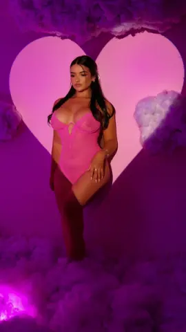 Glamorous woman in a pink bodysuit posing against a dreamy pink heart backdrop with soft cloud decor.