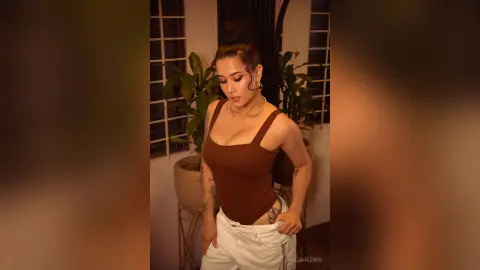 Young woman in a brown bodysuit and white pants posing in a warm, moody indoor setting with plants.