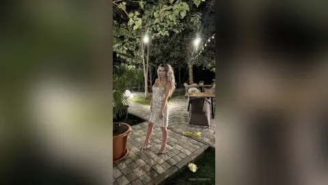 Elegant woman in a sparkling silver cocktail dress standing in a lush outdoor garden patio at night.