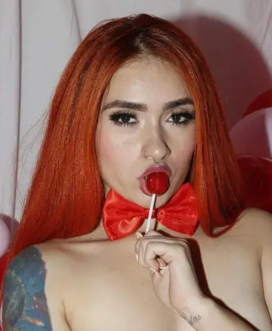 Woman with long red hair holding a red lollipop in a soft pink studio setting.