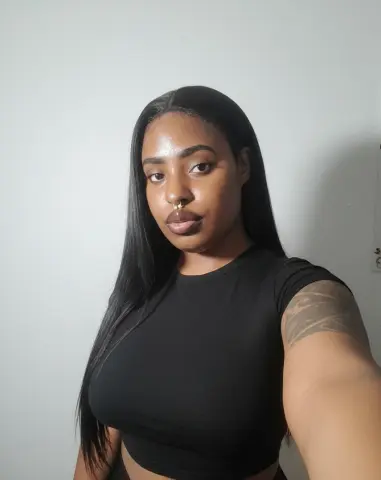 Young Black woman with long straight hair wearing a black t-shirt posing against a plain white wall.