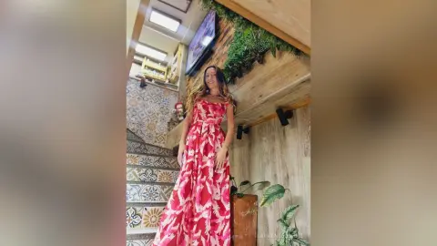 LaurentBelluci stands in a long red and white floral print dress against a light wood wall and tiled stairs.