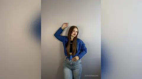 DorisDonachie poses against a gray wall wearing a blue button-down shirt and light blue jeans.