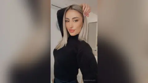 Stylish blonde woman in a black turtleneck sweater posing with a confident expression in a bright room.