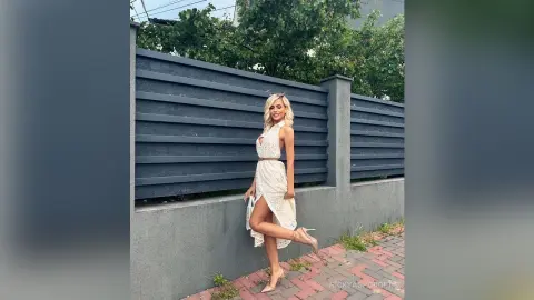 Blonde woman in a white sleeveless maxi dress posing by a modern grey slatted fence on a sunny day.