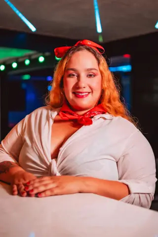 Smiling woman in a white shirt and red vintage headscarf sitting at a neon-lit retro bar.