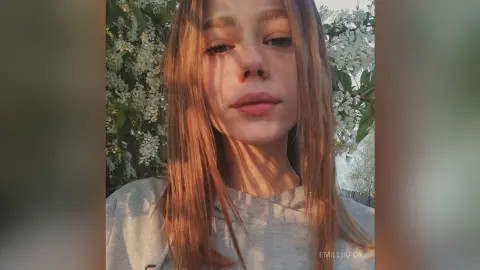 Young woman with long blonde hair wearing a grey sweatshirt in a sunlit garden with white flowers.