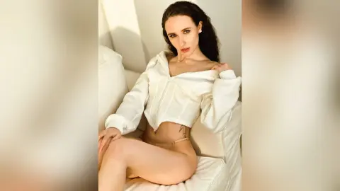 Brunette woman in a white long-sleeve crop top sitting on a white sofa in a bright, minimalist room.