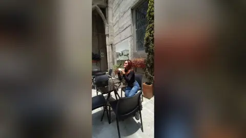 AmelieLautrec sits at a black table outdoors, wearing a black long-sleeved top and blue jeans, holding a white cup or small object near her face.