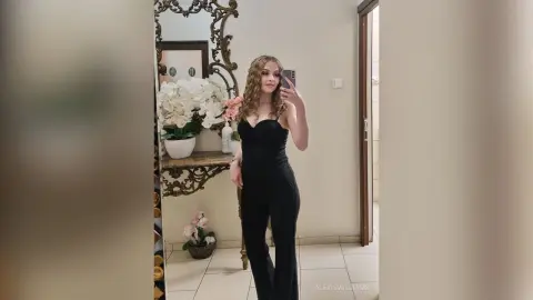 Young woman in a chic black strapless jumpsuit taking a mirror selfie in a bright, modern room.