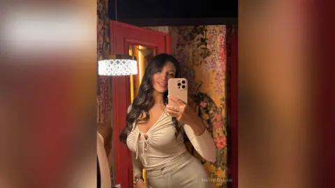 Smiling woman in a beige long-sleeve top taking a mirror selfie in a warm, floral patterned room.