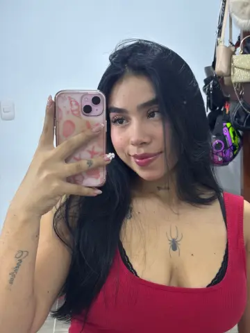 Smiling young woman with long dark hair in a red tank top taking a mirror selfie indoors.