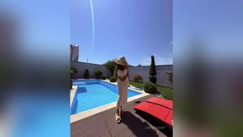Stylish woman in a white summer dress and straw hat relaxing by a bright blue swimming pool on a sunny day.