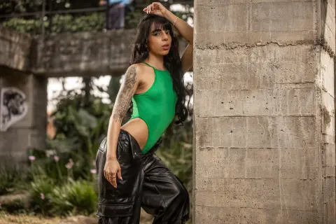 Stylish woman in a green bodysuit and black leather pants leaning against a concrete wall in a lush garden.