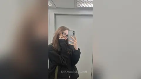 ShakiaGremillion wearing glasses and a black jacket, posing for a mirror selfie with her long brown hair down.