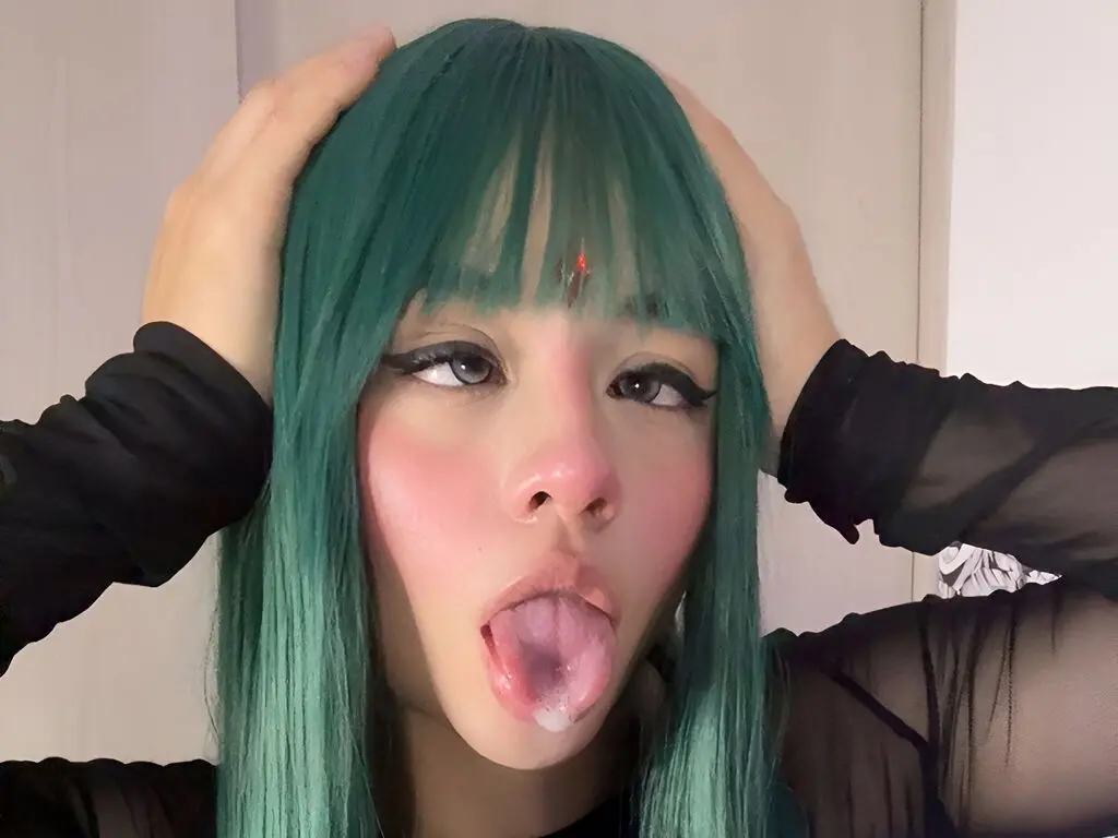 Cosplayer with long teal hair and dramatic eye makeup posing playfully in a black mesh outfit.