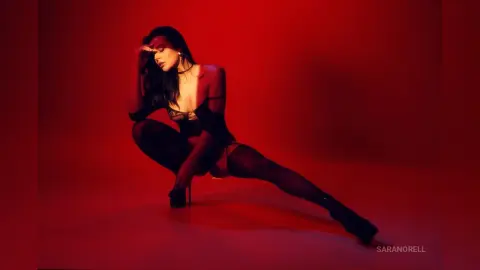 Elegant woman in black lingerie and stockings posing against a moody red studio background.