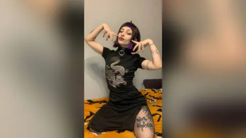 Tattooed woman in a black dragon print dress posing playfully in a bedroom setting.