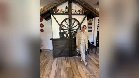 Smiling woman in a beige tracksuit standing in a rustic farmhouse room with a large wooden wagon wheel.
