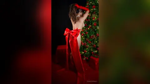 Nude woman wearing a large red satin bow standing in front of a festive Christmas tree in a moody setting.