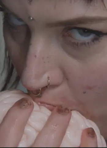 Close-up of a woman with nose piercings biting a soft pink textured object in a moody, intimate setting.