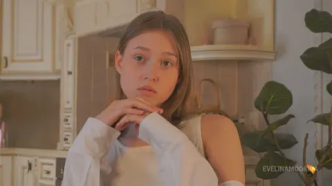 Young woman in a white sleeveless top looking thoughtful in a cozy, sunlit kitchen setting.