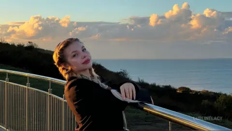 LurlineLacy poses with a smile against a sunset ocean backdrop, leaning on a metal railing with her blonde hair in braids.