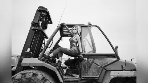 ChristineDavis poses naked in a forklift, sitting on the side with one leg raised and her arms up, wearing black gloves and boots.
