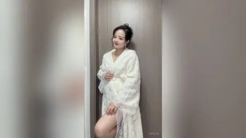 EnyaLin poses in a white fuzzy robe with lace trim, standing against a plain wall.