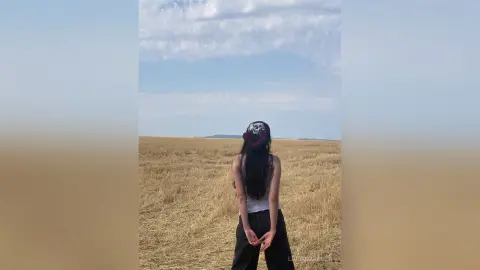 Young woman in a white tank top and black pants standing in a golden wheat field under a cloudy sky.