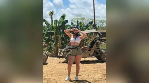 RossiNoleen stands outdoors in a white crop top and olive shorts, holding a black cap over her head against a backdrop of tropical trees.