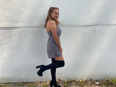 Stylish woman in a black patterned mini dress and over-the-knee boots posing against a white wall.