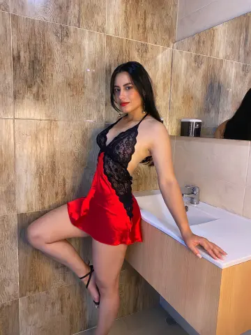 Woman in a red satin and black lace slip dress posing in a modern bathroom with wood-textured tiles.