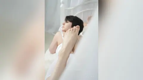 Relaxed woman in white loungewear resting on a bright, airy bed in a peaceful bedroom setting.
